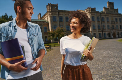 Mirthful young man and woman smiling while walking with notebooks in their hands. Old building on the background